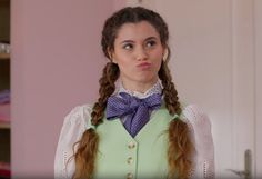 a woman with long hair wearing a green vest and blue bow tie standing in front of a pink wall