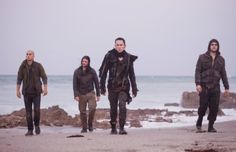 four men walking on the beach in front of the ocean with one wearing a hoodie