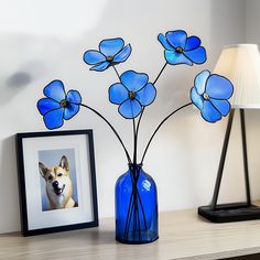 a blue vase with flowers in it next to a framed photo and a lamp on a table