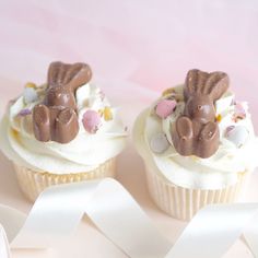 two cupcakes decorated with white frosting and chocolate bunny ears, on a pink surface