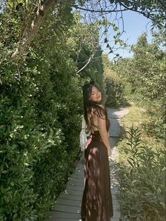 a woman in a long dress standing on a wooden walkway next to bushes and trees