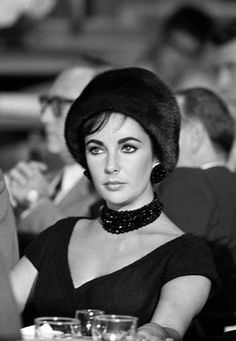 a woman sitting at a table wearing a black dress and hat with pearls on it