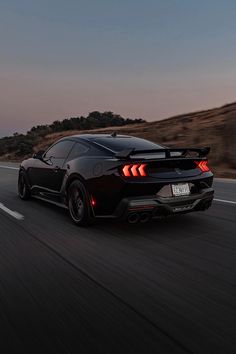 the rear end of a black sports car driving down a road at dusk with hills in the background