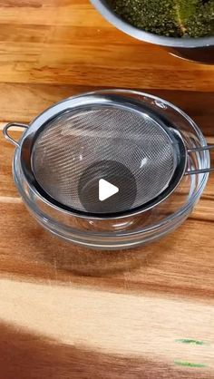 a glass strainer sitting on top of a wooden table next to a bowl filled with broccoli