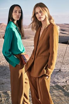 two women standing next to each other in brown pants and green blouses, one with her hand on her hip