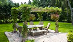 an outdoor dining table and chairs in the middle of a garden with green grass, bushes and trees