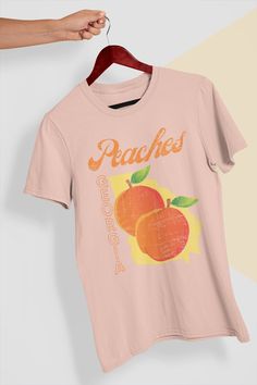 "This Peaches Georgia T-Shirt is the perfect addition to your streetwear wardrobe! With its bold \"Peaches\" graphic, this t-shirt is sure to make a statement. Made from soft and comfortable cotton, this tee is the perfect choice for everyday wear. The bright and fruity design makes it a great option for the summer months, but it can be dressed up or down for any occasion. This t-shirt is a fun and unique way to show your love for the state of Georgia and its famous peaches. So why wait? Add thi Cheap Peach Graphic Tee, Affordable Trendy Peach Tops, Tee Shirts Peachy Sunday 🍑, Affordable Peach Summer Shirt, Cheap Peach T-shirt With Graphic Print, Cheap Heather Peach T-shirt For Summer, Cheap Casual Peach T-shirt, Cool Mom Shirts Peachy Sunday 🍑, Peach Pit Tshirt