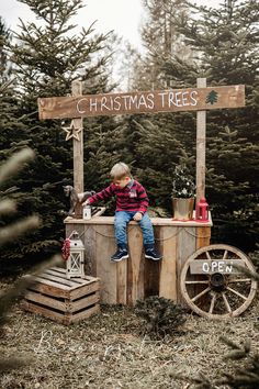 a young boy sitting on top of a wooden box in front of christmas tree stand