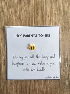 a bee pin sitting on top of a wooden table next to a sign that says, hey parents - to - bee wishing you all the honey and happiness as you welcome your little bee