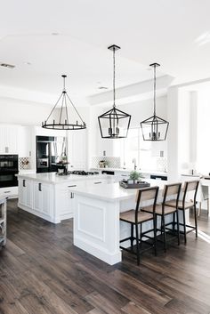 a large kitchen with white cabinets and wooden flooring is pictured in this image, there are several hanging lights above the island