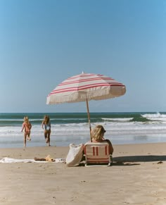 two women are sitting under an umbrella on the beach while another woman is running in the water