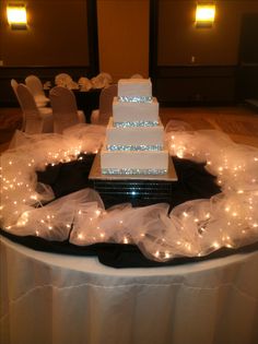 a white wedding cake sitting on top of a table covered in tulle and lights