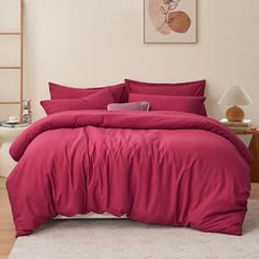 a bed with red sheets and pillows in a white room next to a painting on the wall