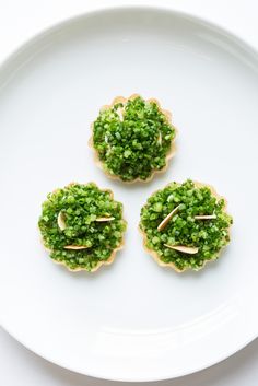 three small cookies with green toppings on top of a white plate, ready to be eaten