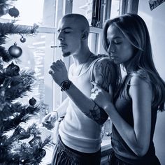 a man and woman standing next to a christmas tree with candles in their hands looking at each other