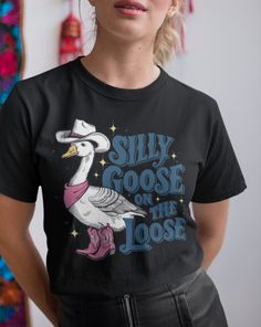 Get ready to turn heads and spread smiles with our "Silly Goose on the Loose" T-shirt! This playful design features an adorable goose dressed to impress in a cowboy hat, bandana, and pink cowboy boots, perfectly capturing a whimsical Western vibe. Whether you're a fan of quirky animal graphics or looking for a fun gift, this tee is sure to delight. Product Details:     Material: Made from soft, high-quality cotton for all-day comfort     Design: Unique hand-drawn graphic of a goose in Western attire, complete with fun typography     Fit: Available in various sizes, from XS to 5XL, with a relaxed, unisex fit     Colors: Choose from a range of vibrant colors to suit your style Novelty Cotton Top With Sublimation Print, Novelty Cotton Tops With Sublimation Print, Novelty Cotton Shirt With Screen Print, Novelty Cotton Tops With Screen Print, Novelty Cotton Shirt Pre-shrunk, Goose Dress, Cute Goose, Pink Cowboy Boots, Fun Typography