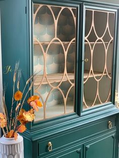 a vase with flowers in it sitting next to a green china cabinet on top of a wooden floor