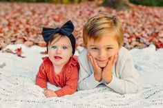 two young children laying on top of a white blanket in the fall leaves with their hands under their chins