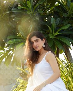 a beautiful young woman in a white dress sitting on a bench next to some trees