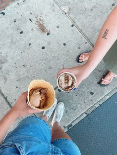 two people holding cups of coffee and ice cream on the sidewalk with their feet crossed