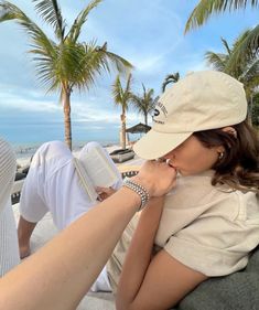 a woman reading a book while sitting on a bench next to the ocean and palm trees