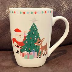 a white coffee cup with santa and reindeer around it on a brown leather surface next to a christmas tree