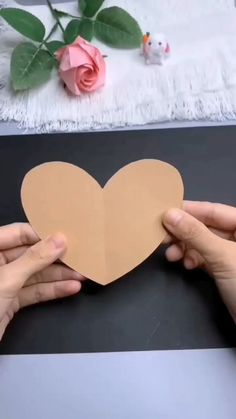 two hands holding up a paper heart on a table next to a rose and a vase