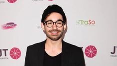 a man wearing glasses and a black hat smiles at the camera while standing in front of a pink backdrop