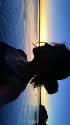 a woman standing next to the ocean with her reflection in the water and sun setting behind her