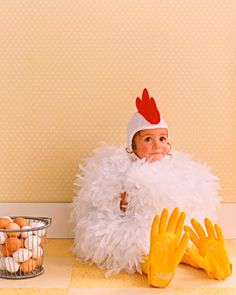 a baby dressed up as a chicken sitting on the floor next to eggs and gloves