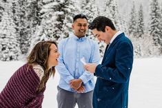 two men and a woman standing in the snow talking to each other with trees in the background