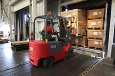 a man driving a red forklift in a warehouse with boxes on the floor and another person behind it