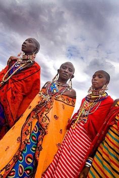 three african women wearing colorful clothing and headdress standing in front of a cloudy sky