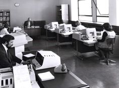 black and white photograph of people working on computers in an office with many desks