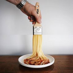 a hand holding a wooden spatula filled with spaghetti on top of a white plate