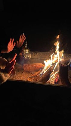 two people sitting at a campfire with their hands up in the air above them