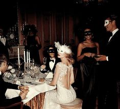 a group of people sitting around a dinner table with masks on their faces and one woman in a white dress