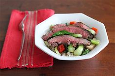 a white bowl filled with meat and vegetables on top of a wooden table next to a red napkin