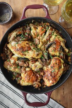 a pan filled with chicken and mushrooms on top of a wooden table next to wine glasses