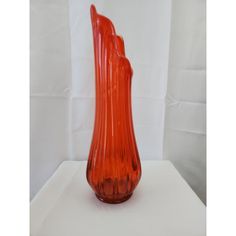 an orange glass vase sitting on top of a white tableclothed backdrop with the image of a tall, slender figure behind it