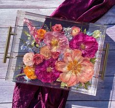 an arrangement of flowers in a clear box on a purple velvet cloth and wood background