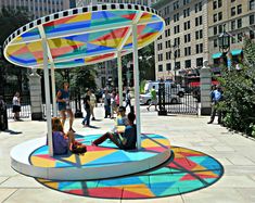 several people are sitting on the ground under a colorfully painted umbrella in a city square