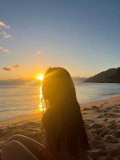 a woman sitting on top of a sandy beach next to the ocean at sun set