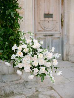 some white flowers are sitting in a vase on the ground near a door and shrubbery