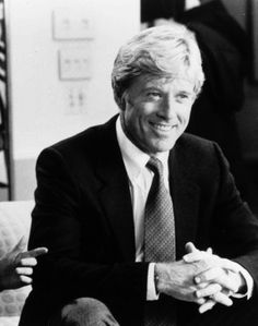 a man in a suit and tie sitting on a couch with his arms crossed, smiling