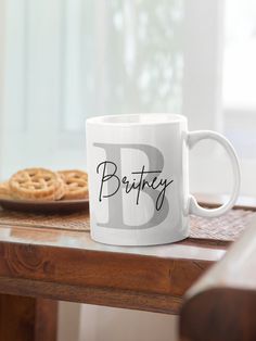 a white coffee mug sitting on top of a wooden table next to a plate of cookies