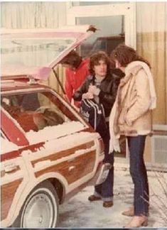 two women standing next to a car with the hood open and it is covered in snow