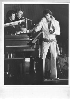 elvis presley singing into a microphone while standing in front of a piano and another man behind him