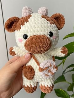 a hand holding a brown and white crocheted stuffed animal next to a potted plant