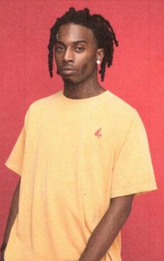 a young man with dreadlocks standing in front of a red wall wearing a yellow shirt